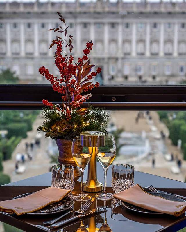 Mesa con un centro navideño de bayas rojas y dos copas de vino, con el Palacio Real al fondo